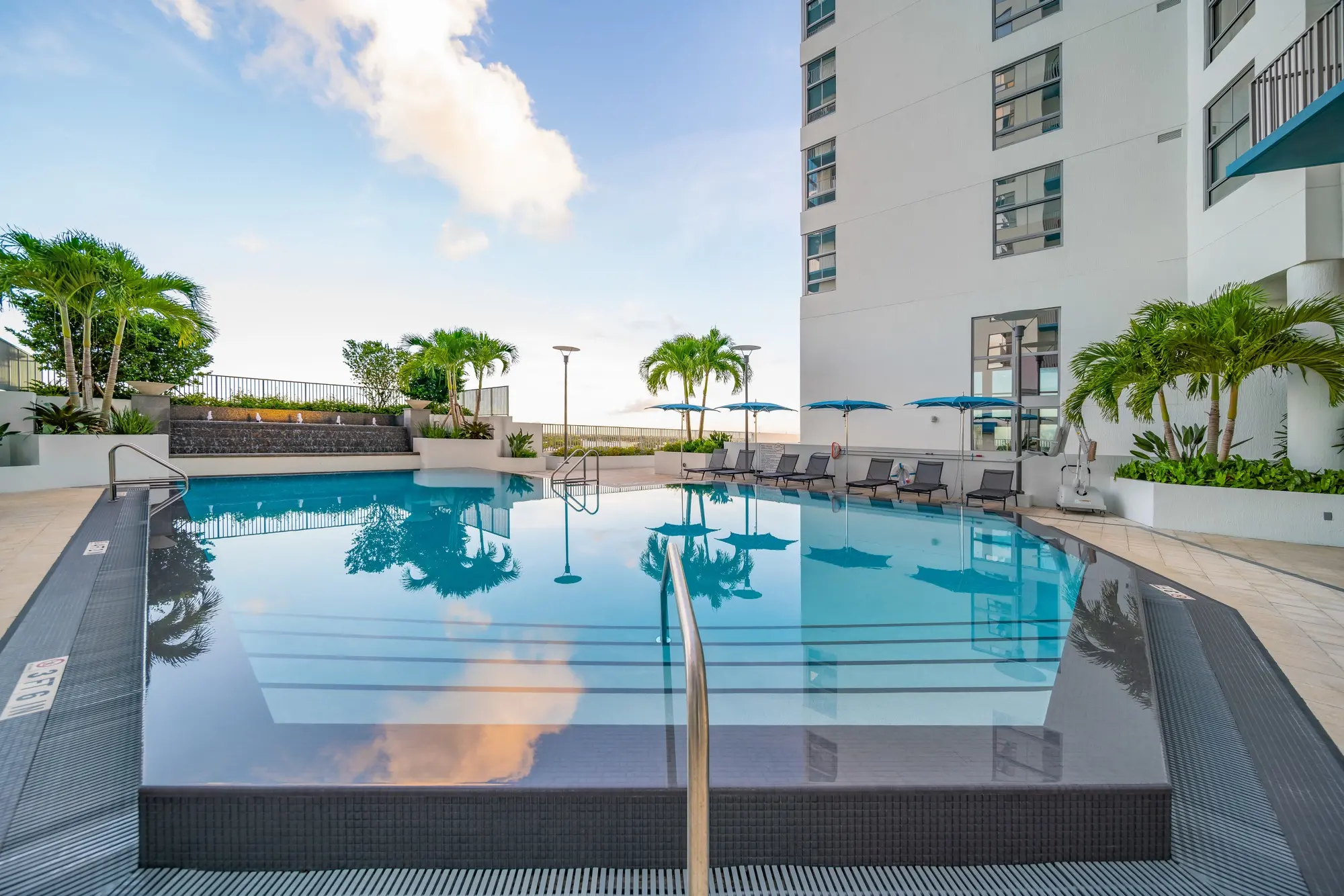 A modern swimming pool surrounded by palm trees and lounge chairs, with a high-rise building in the background and a clear blue sky.