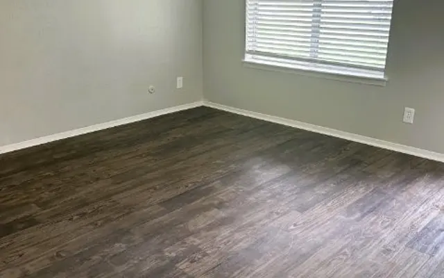 Empty room with light-colored walls, a window with blinds, and wooden flooring. Natural light brightens the space.