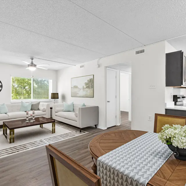 A modern apartment interior showcasing a living room with a grey couch, brown coffee table, and dining area with a round table and chairs, featuring light-colored walls and flooring, and bright natural light coming through large windows.