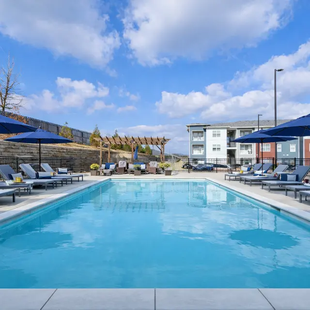 A beautifully designed swimming pool surrounded by lounge chairs and umbrellas, with an apartment complex in the background and a clear blue sky.