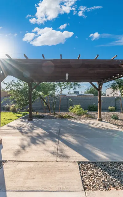 Wooden Pergola in Backyard A wooden pergola in a backyard with a concrete patio, surrounded by grass and desert vegetation, under a blue sky with clouds.