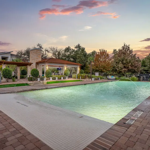 A serene outdoor swimming pool area featuring a large pool with a gentle gradient, surrounded by trees and landscaping, under a colorful sunset sky.