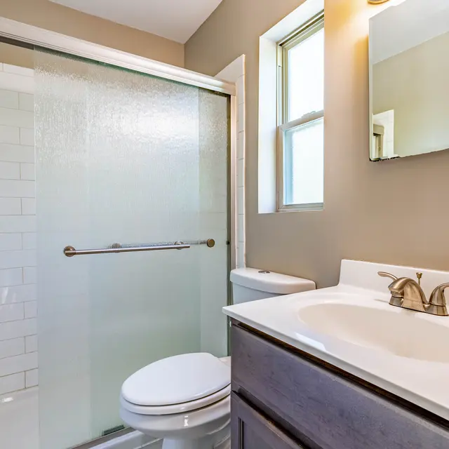 A modern bathroom featuring a glass walk-in shower, a white countertop basin with a sink, and a toilet. The walls are a warm beige color, and there's a rectangular mirror above the sink. Natural light enters through a window next to the shower.