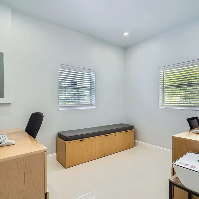 A modern office interior featuring two desks, a seating area, and large windows with blinds. The walls are painted light blue, and the flooring looks glossy. One desk has a computer and a chair, while the other has a printer. The space is well-lit and minimalistic.