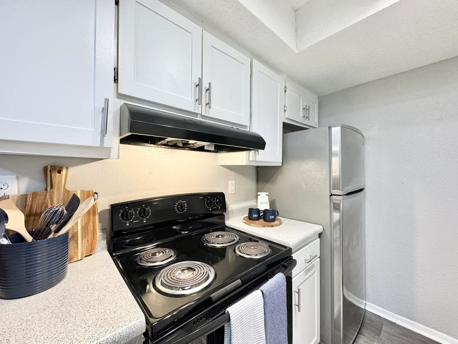 A modern kitchen featuring a black stove, white cabinetry, and stainless steel refrigerator. Utensils are neatly arranged on a countertop along with decorative items.