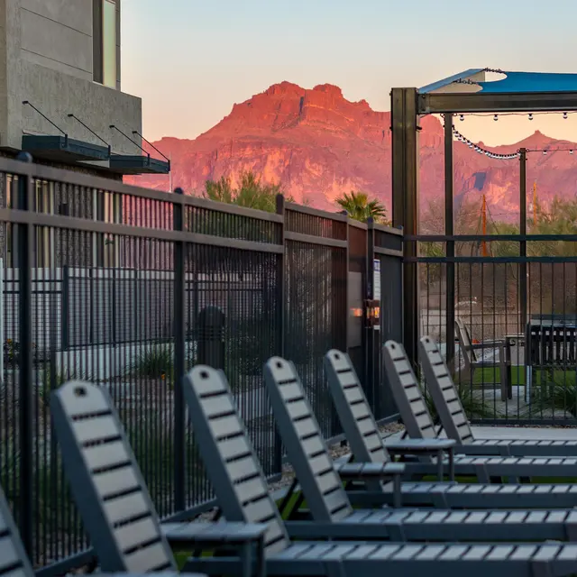 An outdoor pool area featuring several lounge chairs in the foreground, with a backdrop of a mountain illuminated by the sunset.