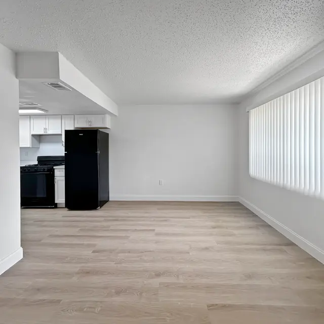 Modern Apartment Interior A view of an empty modern apartment interior featuring a light wooden floor, a kitchen area with white cabinets and a black refrigerator. Large window with vertical blinds allows natural light into the space.