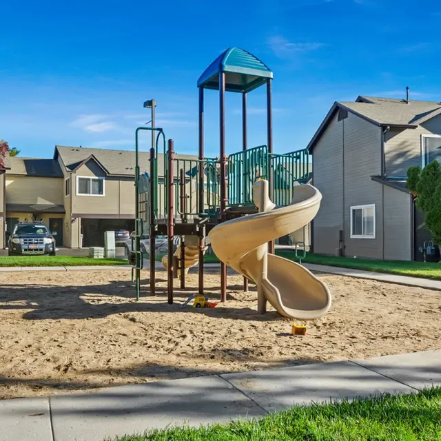 Monterra Townhomes - Playground with Slide A playground featuring a tan slide on a sandy ground, surrounded by grassy areas and residential buildings in the background.