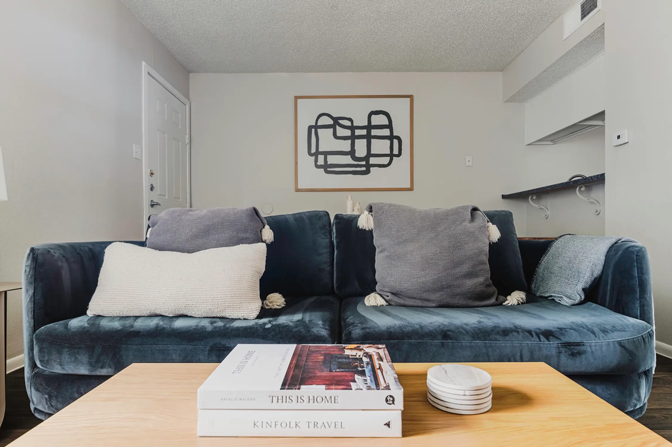 A cozy living room featuring a plush blue couch with two decorative pillows and a wooden coffee table. On the table, there are stacked books and small white plates. A minimalist artwork is hung on the wall.
