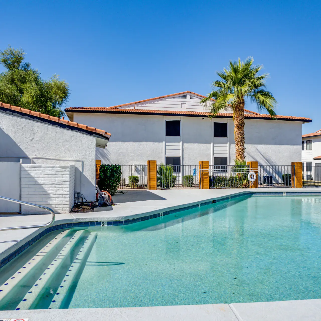 A clear swimming pool surrounded by a white building with a red-tiled roof and palm trees in the background under a bright blue sky.