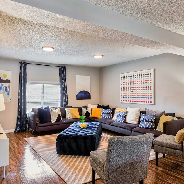 A stylish modern living room with a sectional sofa, colorful decorative pillows, and a large coffee table. The walls are adorned with abstract art and the space features two lamps and textured flooring.