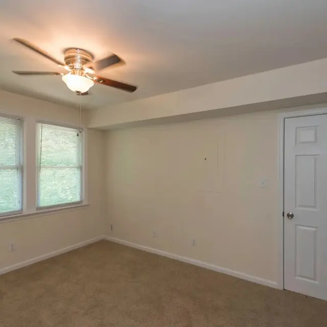 An empty room featuring beige walls, carpeted floor, a ceiling fan, and two windows with blinds.