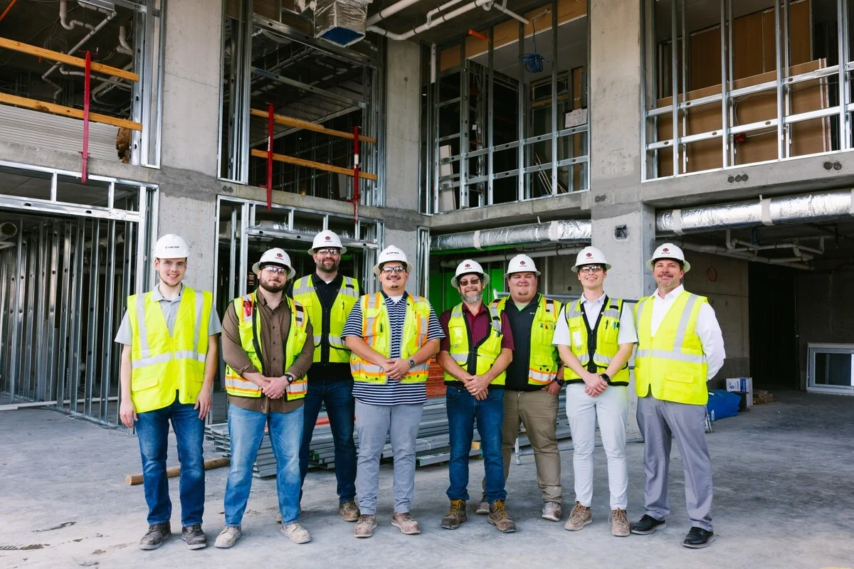A group of eight construction professionals standing together on a construction site. They are wearing hard hats and high-visibility vests, with a partially completed building in the background.