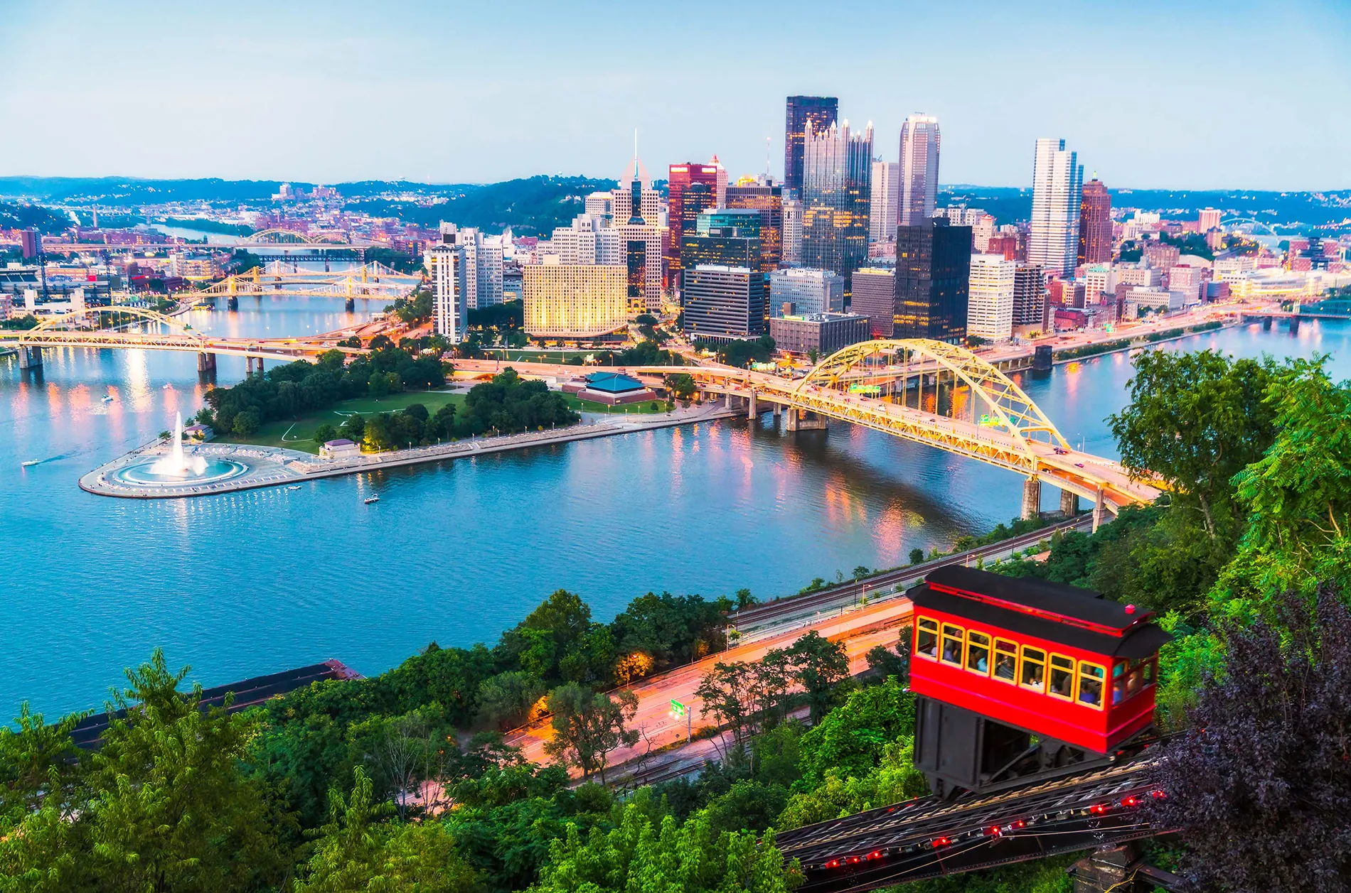 Pittsburgh Skyline at Sunset A panoramic view of Pittsburgh, Pennsylvania, showcasing tall buildings, a river, and a red funicular railway car descending the hill.