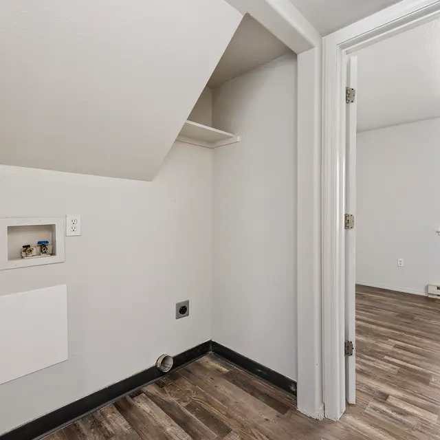 Room Interior with Alcove A photo of a corner of a room featuring a small alcove with a white wall and a shelf above. There is a panel on the wall, and the floor is made of wooden planks. A doorway leads to another area of the room, which has a large window letting in natural light.
