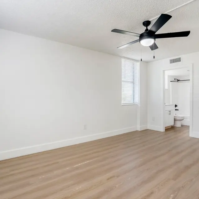Empty Room with Ceiling Fan and Wood Flooring A spacious, empty room with light-colored wooden flooring, white walls, and a ceiling fan. A window with blinds allows natural light in. A doorway leads to an adjacent bathroom space.