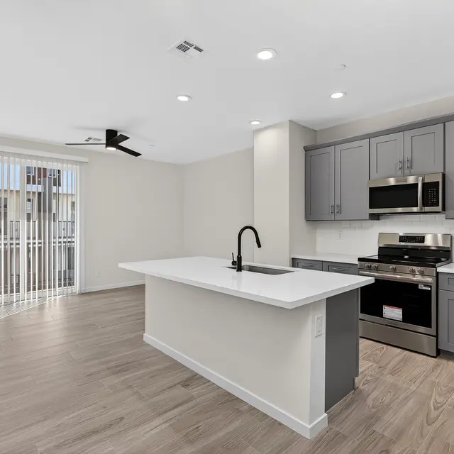 A modern kitchen in an apartment, featuring a central island with a sink, stainless steel appliances, and dark cabinetry. Large windows with vertical blinds provide natural light, and the flooring is wood-like.