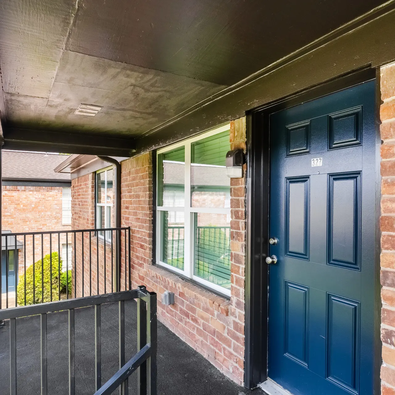 A close-up view of an apartment entryway featuring a dark blue door framed by brick walls and a small covered porch area with metal railings.