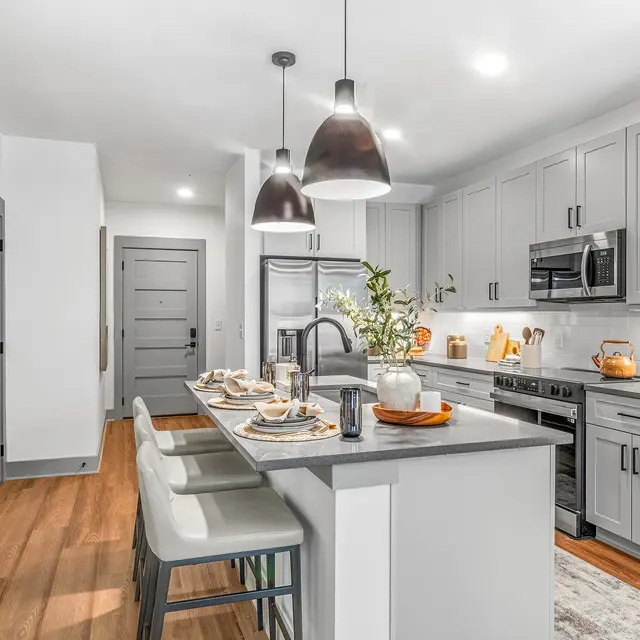 Modern Kitchen Interior A modern kitchen featuring gray cabinets, stainless steel appliances, and a large island with seating. Pendant lights hang above the island, which is set with tableware and decor. The flooring is hardwood, and the walls are painted white.