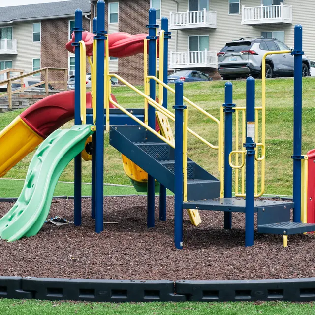 Colorful playground equipment featuring slides, climbing structures, and steps, situated on grass near apartment buildings.