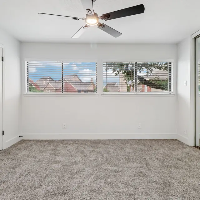 A spacious bedroom with light gray carpet, featuring a ceiling fan and large windows showing a view of houses outside.