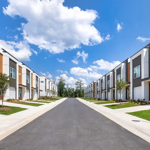 Street view of modern residential buildings lined with greenery under a blue sky with clouds.