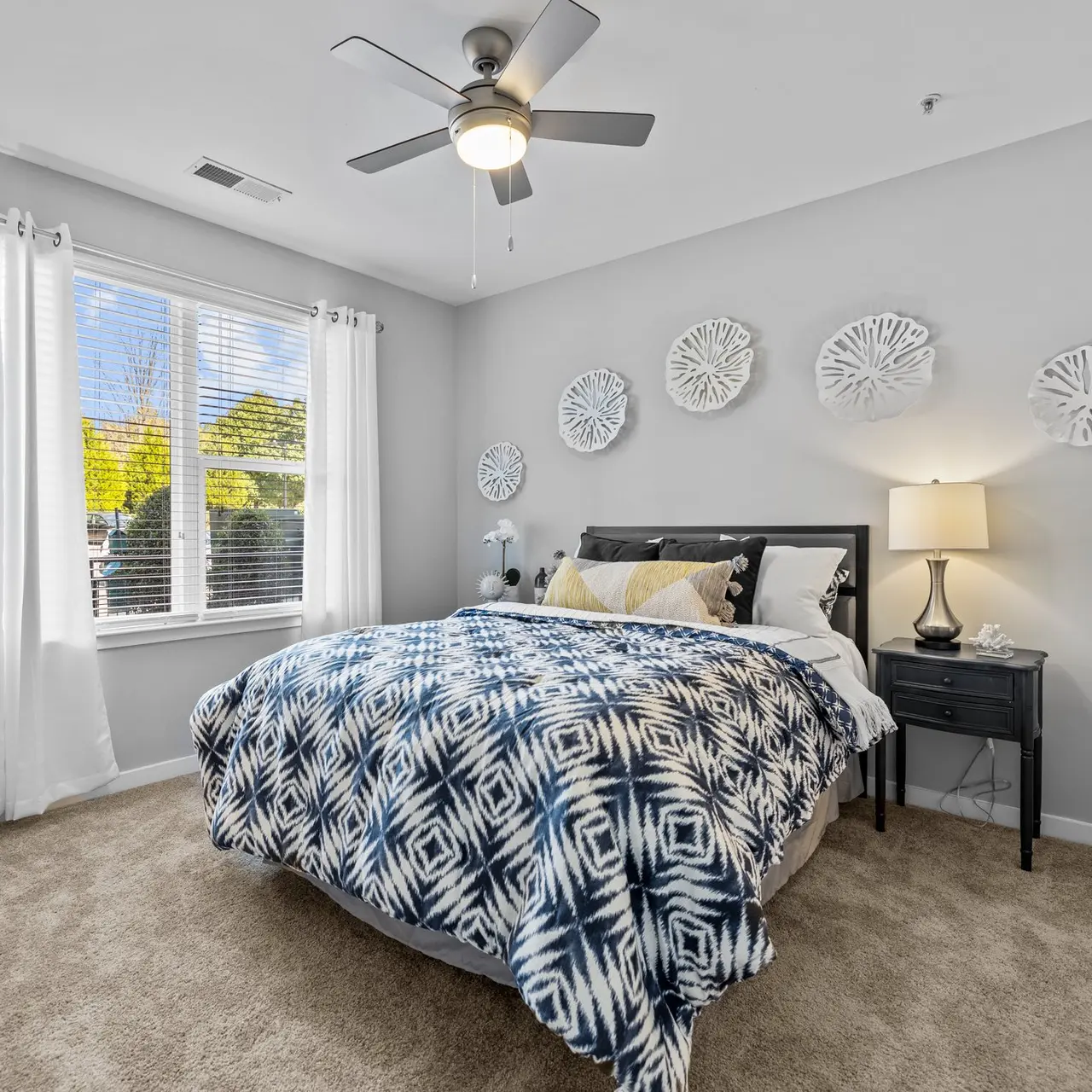 A cozy bedroom featuring a queen-sized bed with a patterned blue and white blanket, a nightstand with a lamp, and large windows with white curtains.