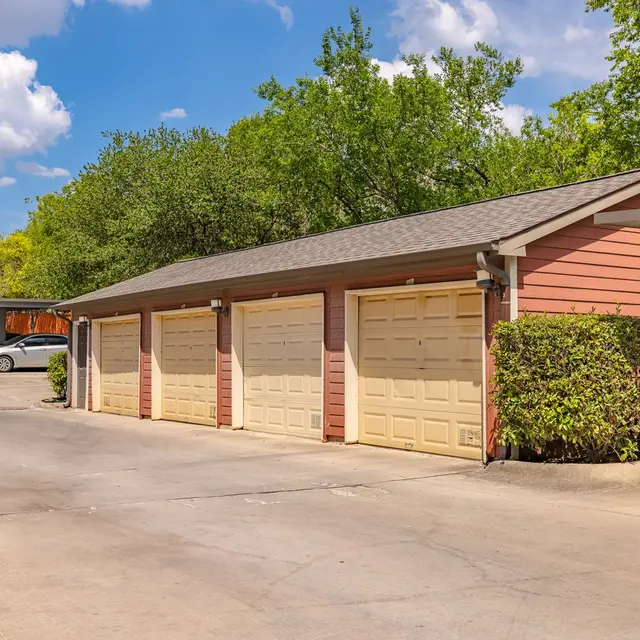 A row of beige garage doors attached to a red building, surrounded by green foliage under a bright blue sky with fluffy white clouds.