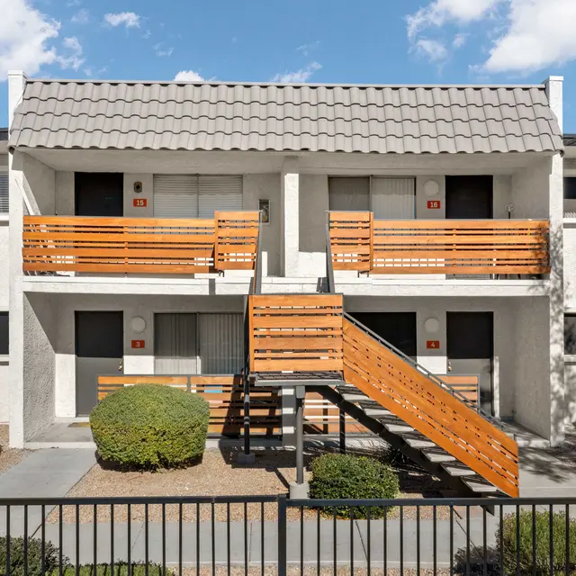 A two-story apartment building featuring a tiled roof, orange wooden balconies, and an exterior staircase. The ground is landscaped with small shrubs and gravel.