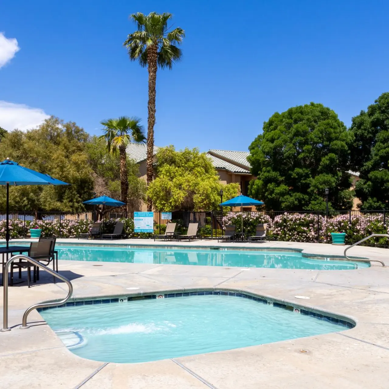 A well-maintained outdoor pool area featuring a large swimming pool, a hot tub, several blue umbrellas, and lounge chairs, surrounded by lush greenery and palm trees under a bright blue sky.