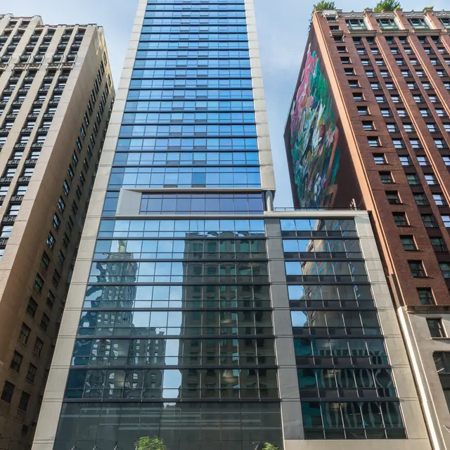 A tall modern building with a glass facade, surrounded by other high-rise structures. The building has a red accent at the top and features greenery at its base.