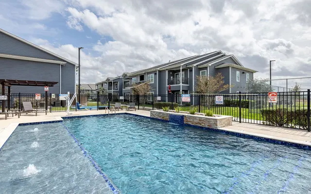 A swimming pool surrounded by a patio and lounge chairs, with an apartment complex in the background and partly cloudy skies above.