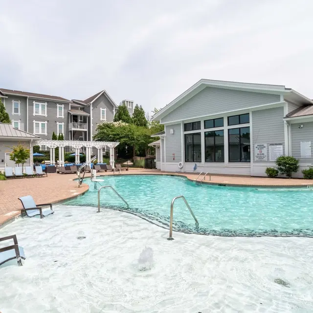 A spacious outdoor swimming pool area with lounge chairs and a modern building in the background. The pool has a hot tub section integrated into it.