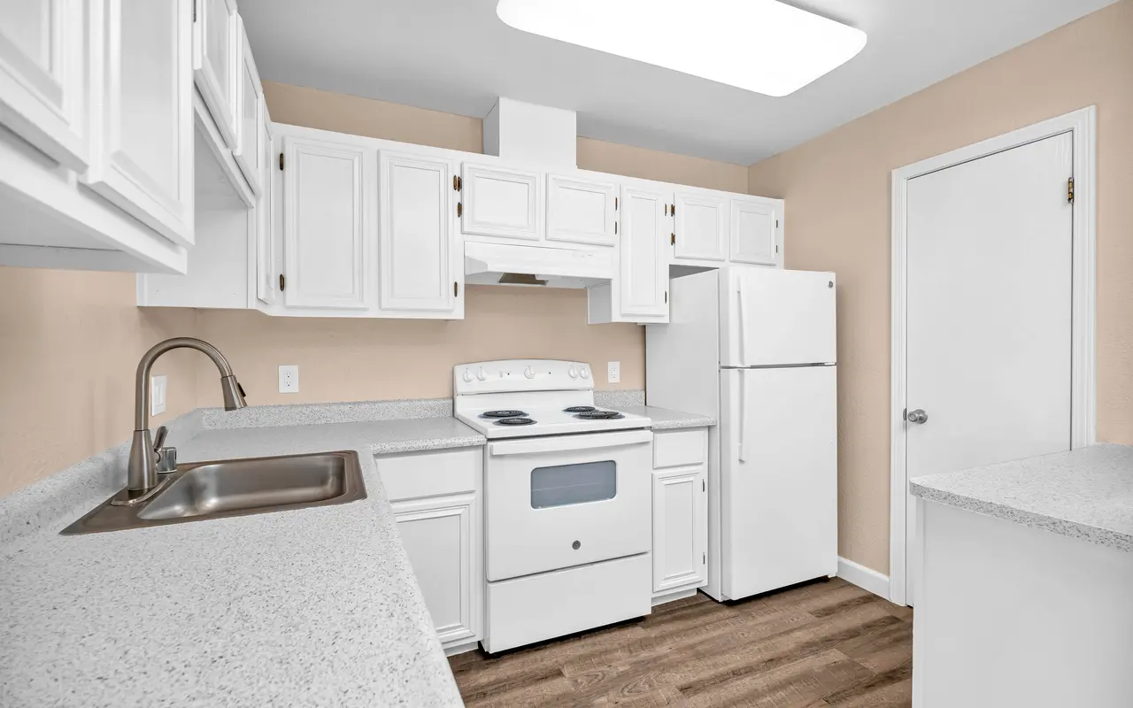 A modern kitchen with white cabinetry, a stainless steel sink, and appliances including a stove and refrigerator, set against a light beige wall.