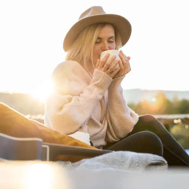 A woman sitting outdoors holding a cup, wearing a hat and a cozy sweater. The sun is setting in the background, creating a warm glow.