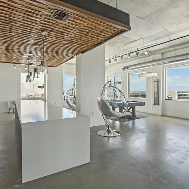A spacious modern office interior featuring a white countertop, wood-paneled ceiling, and large windows with a view. There are contemporary chairs and a hanging chair in the layout.