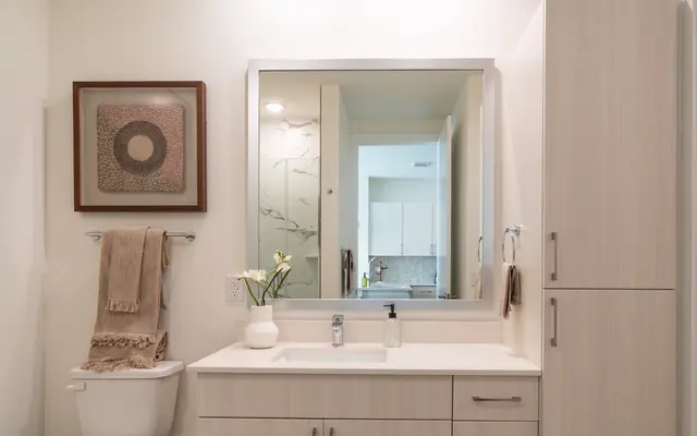Modern Bathroom Interior A modern bathroom featuring a wooden floor, a white vanity with a sink, a toilet, and a tall storage cabinet. The wall is adorned with a framed artwork and a mirror reflecting the interior. Soft lighting illuminates the space.