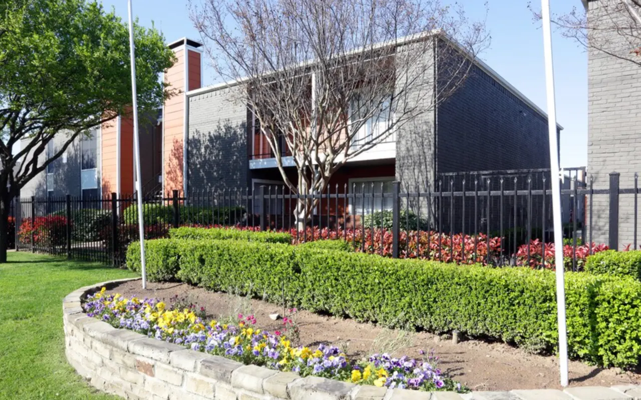 An exterior view of a building surrounded by green grass and flower beds featuring colorful flowers. There are trees nearby and a black fence along the perimeter.