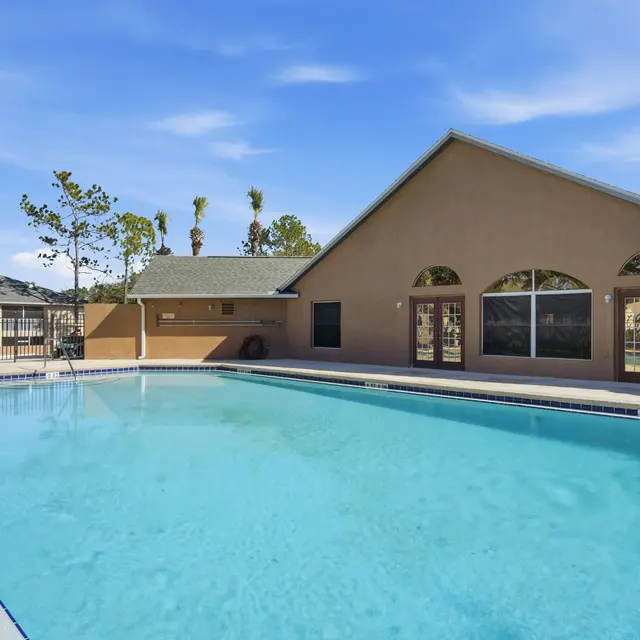 A clear blue swimming pool next to a tan building with large windows, surrounded by trees and a fence.