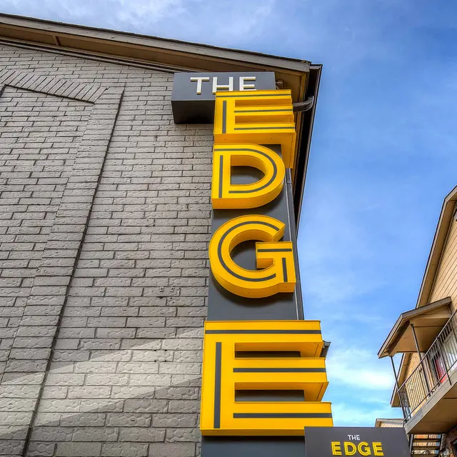 Close-up of a sign that reads 'THE EDGE' in bold yellow letters against a gray wall with blue sky in the background.
