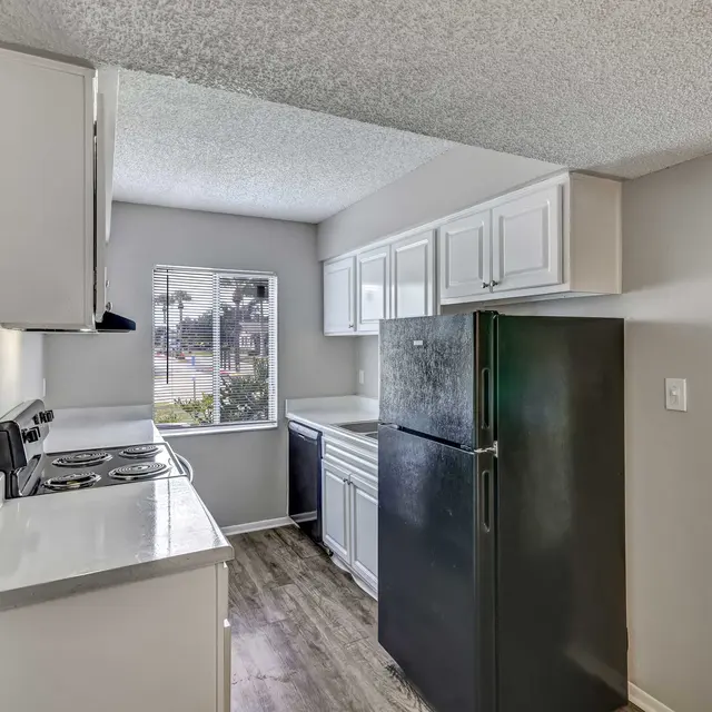 A modern kitchen with white cabinets, a black refrigerator, and a window showing a view outside. The kitchen features a stove and a light-colored countertop, with wood-like flooring.