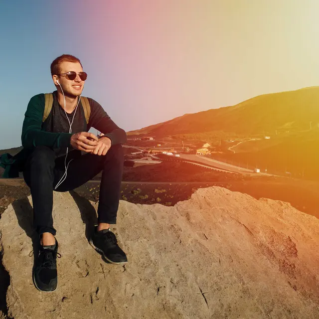 A young man sitting on a rock against a mountainous backdrop, wearing sunglasses and headphones while holding a phone.
