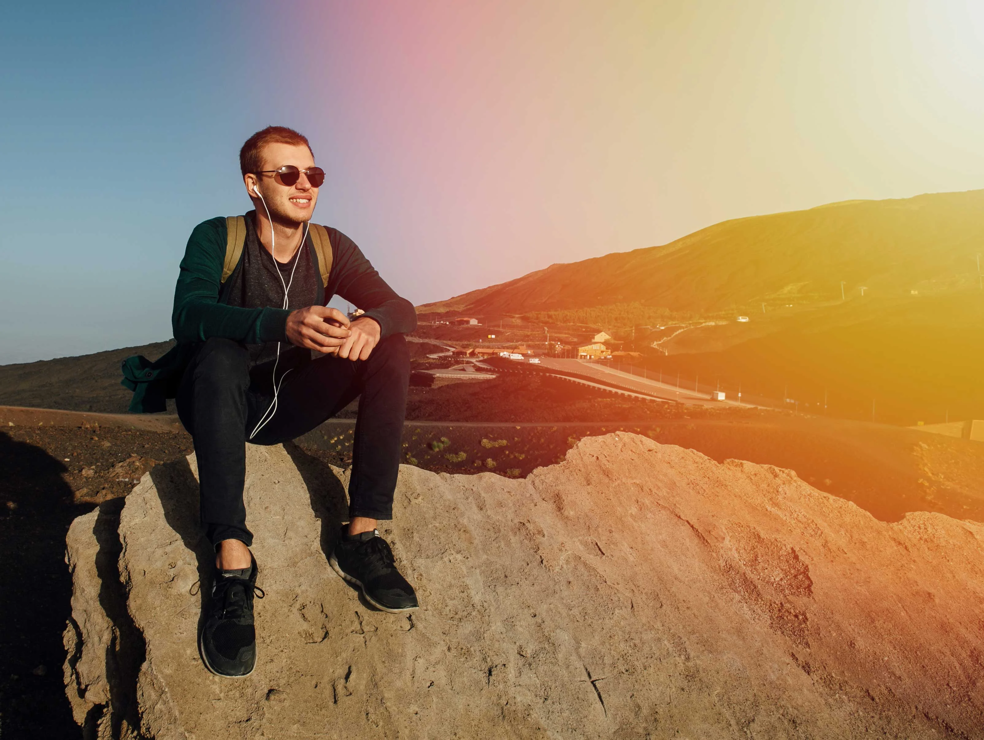 A young man sitting on a rock against a mountainous backdrop, wearing sunglasses and headphones while holding a phone.