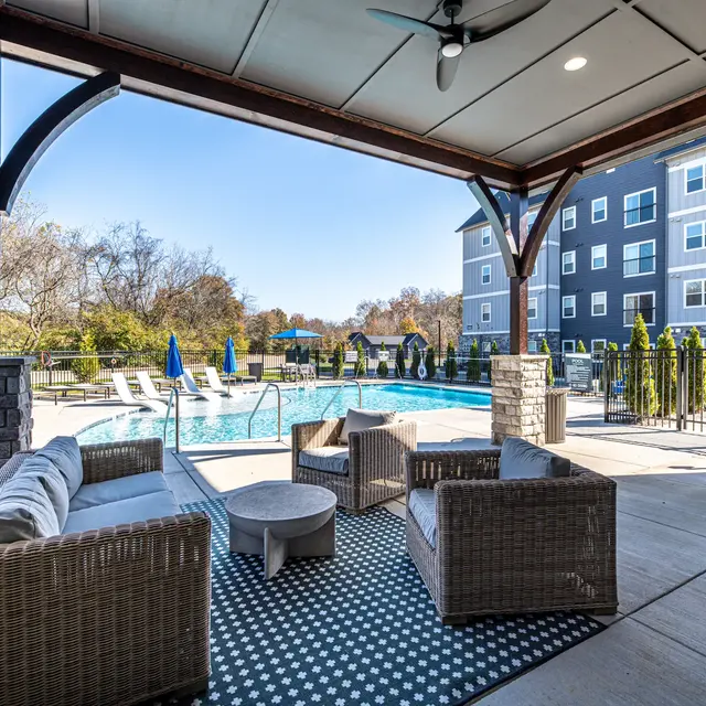 A cozy outdoor lounge area with wicker furniture under a covered patio, overlooking a swimming pool surrounded by trees and tall buildings.