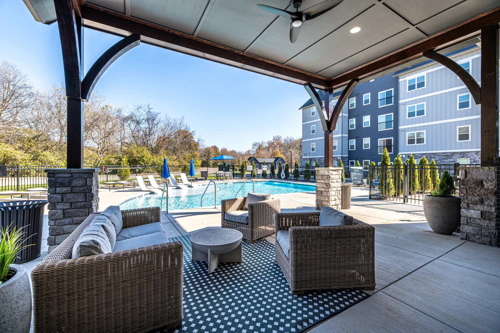 A cozy outdoor lounge area with wicker furniture under a covered patio, overlooking a swimming pool surrounded by trees and tall buildings.