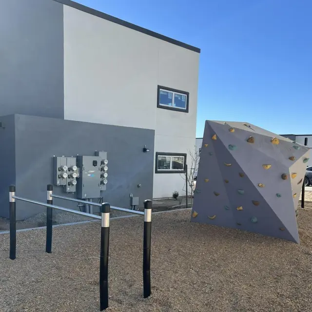 A modern climbing structure outside a building, featuring a triangular bouldering wall in gray with colorful holds and a set of exercise bars nearby.