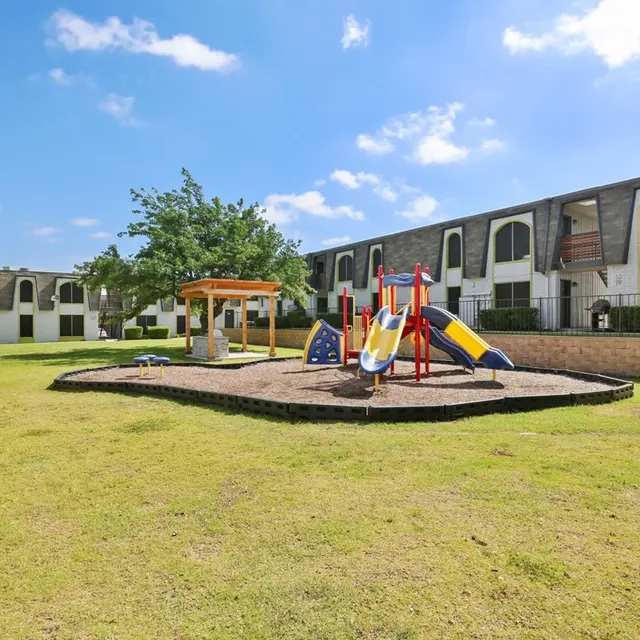 A colorful playground with slides and climbing structures in a grassy area surrounded by apartment buildings under a blue sky.