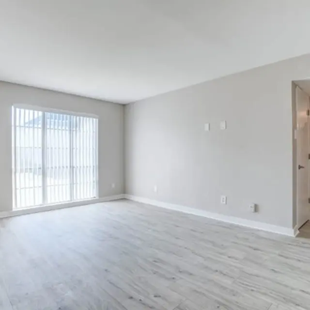 A spacious, empty living room with light-colored wooden flooring and a large window with blinds. The walls are painted in a light neutral tone, and there are doors leading to other rooms visible in the background.