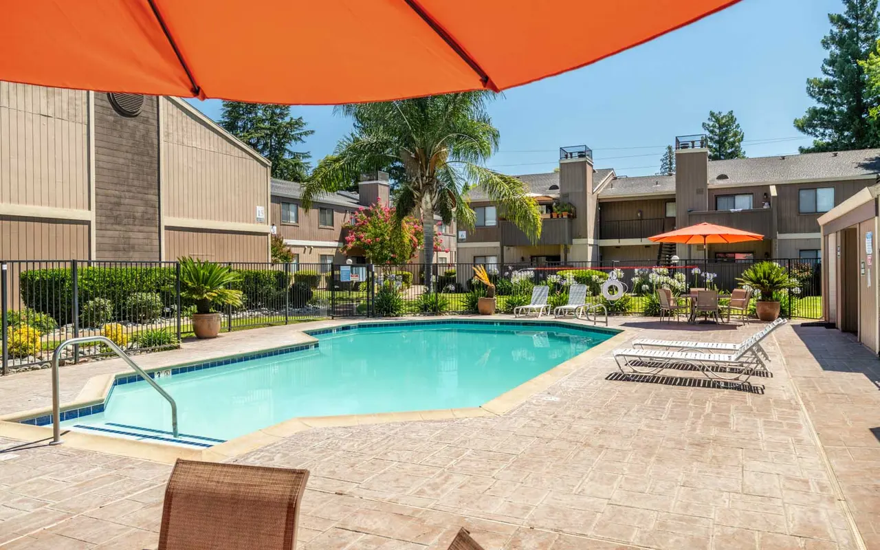 A bright and inviting apartment pool area with a clean swimming pool surrounded by lounge chairs and tropical plants. There are umbrellas providing shade, and a backdrop of two-story apartment buildings under a clear blue sky.
