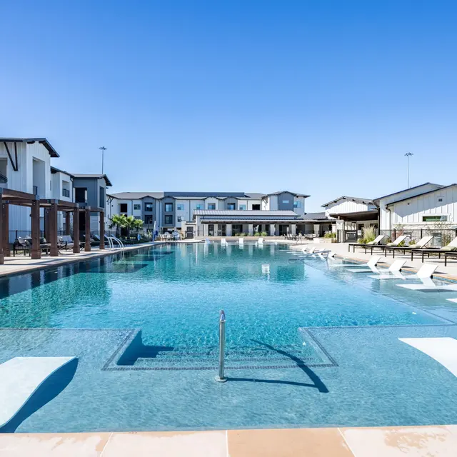 A luxury apartment complex featuring a large swimming pool surrounded by lounge chairs and modern architecture in the background.